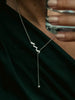 A hero close-up of a person wearing a delicate silver Y-necklace with a zigzag pendant and a dangling chain ending in a small bead, styled over a dark green top.